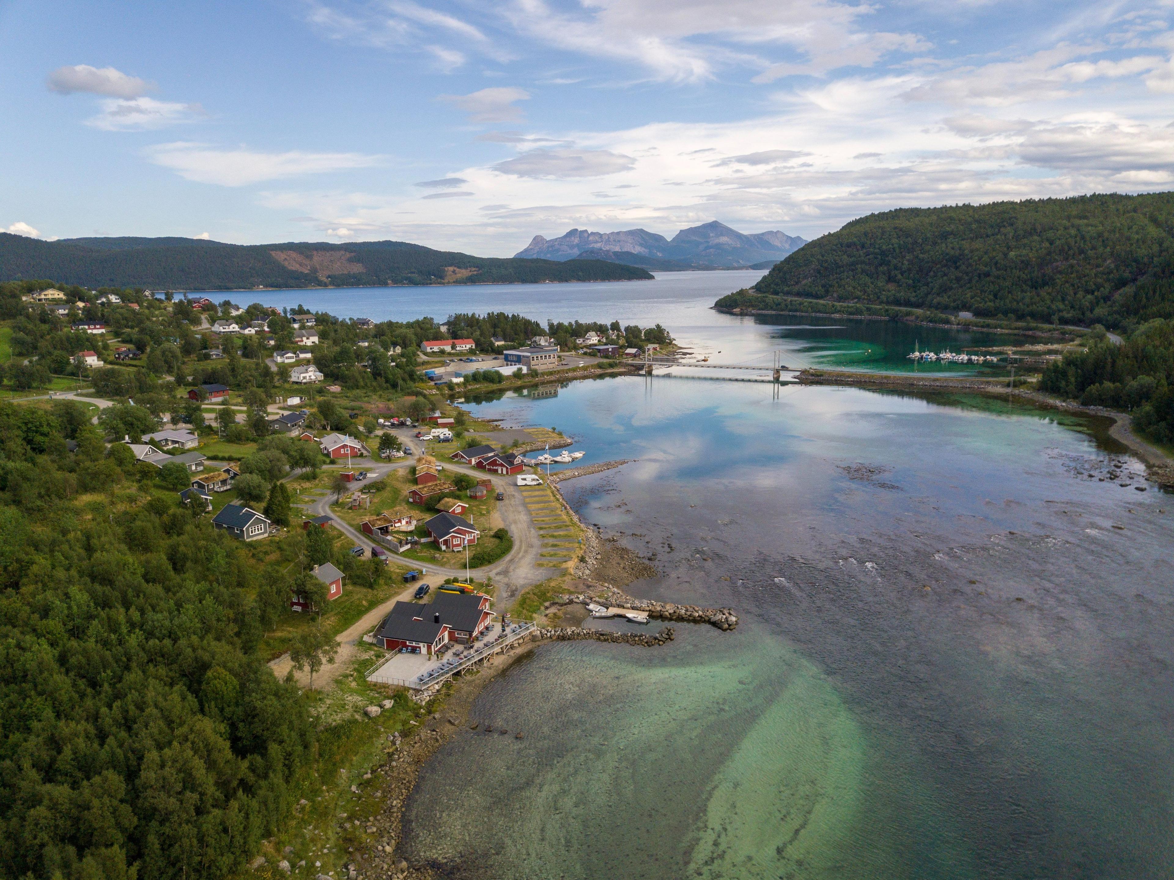 Luftbilde av en vakker kystlinje med hus, fjord og fjell i bakgrunnen. Klar blå himmel og rolig vann.