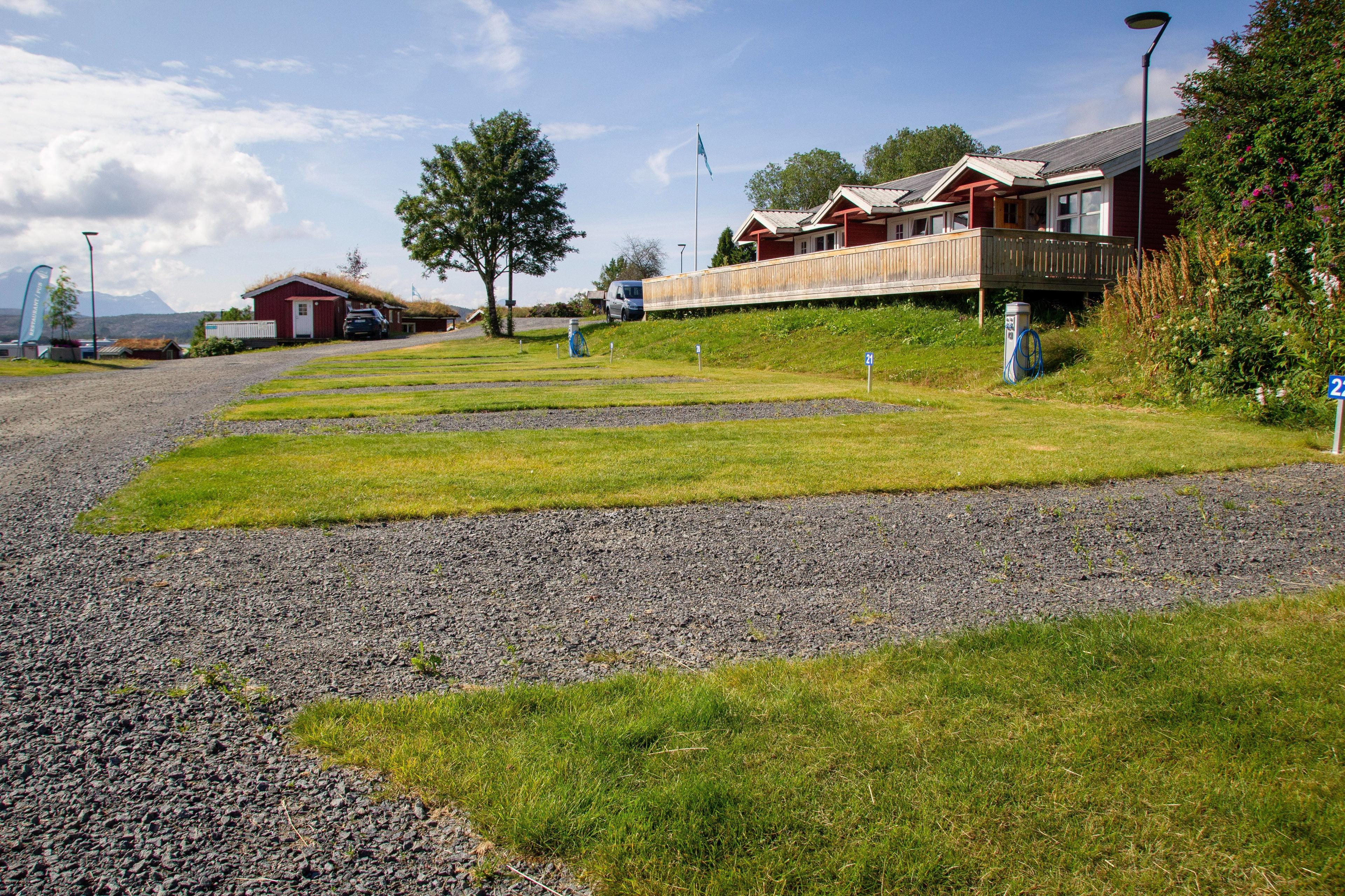 Utsikt over campingplass med grusveier, gressplener og røde hytter i bakgrunnen under en blå himmel.