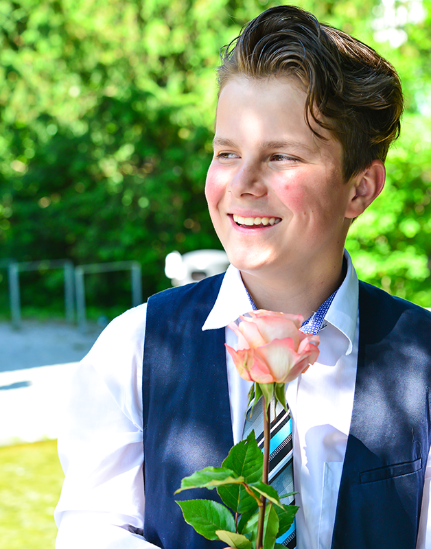 En smilende gutt i dress holder en rosa rose, med grønn bakgrunn av trær.