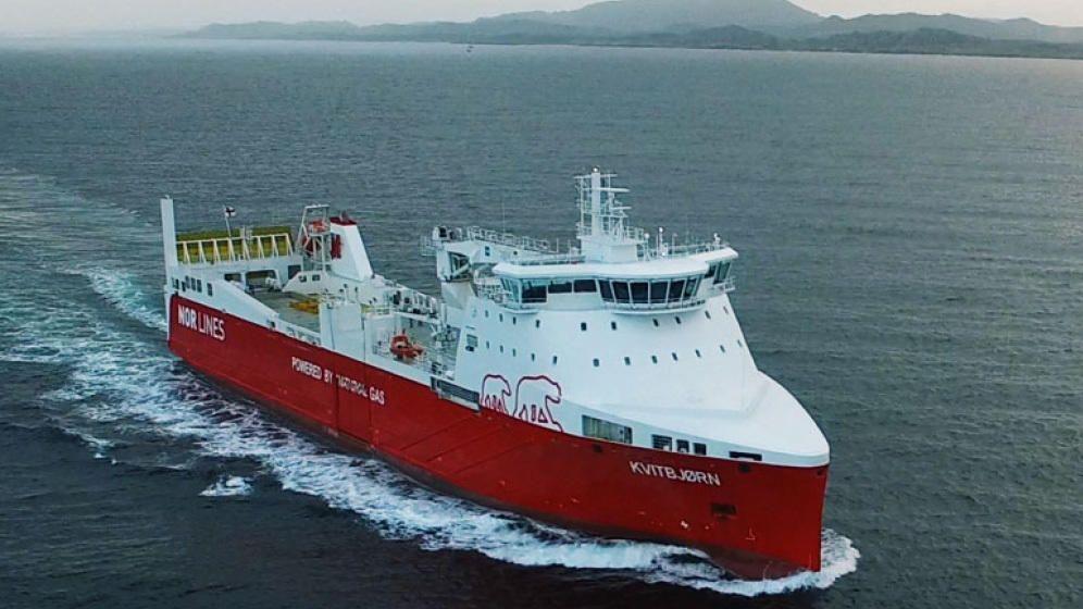Et stort rødt fraktskip seiler på havet med fjell i bakgrunnen.
