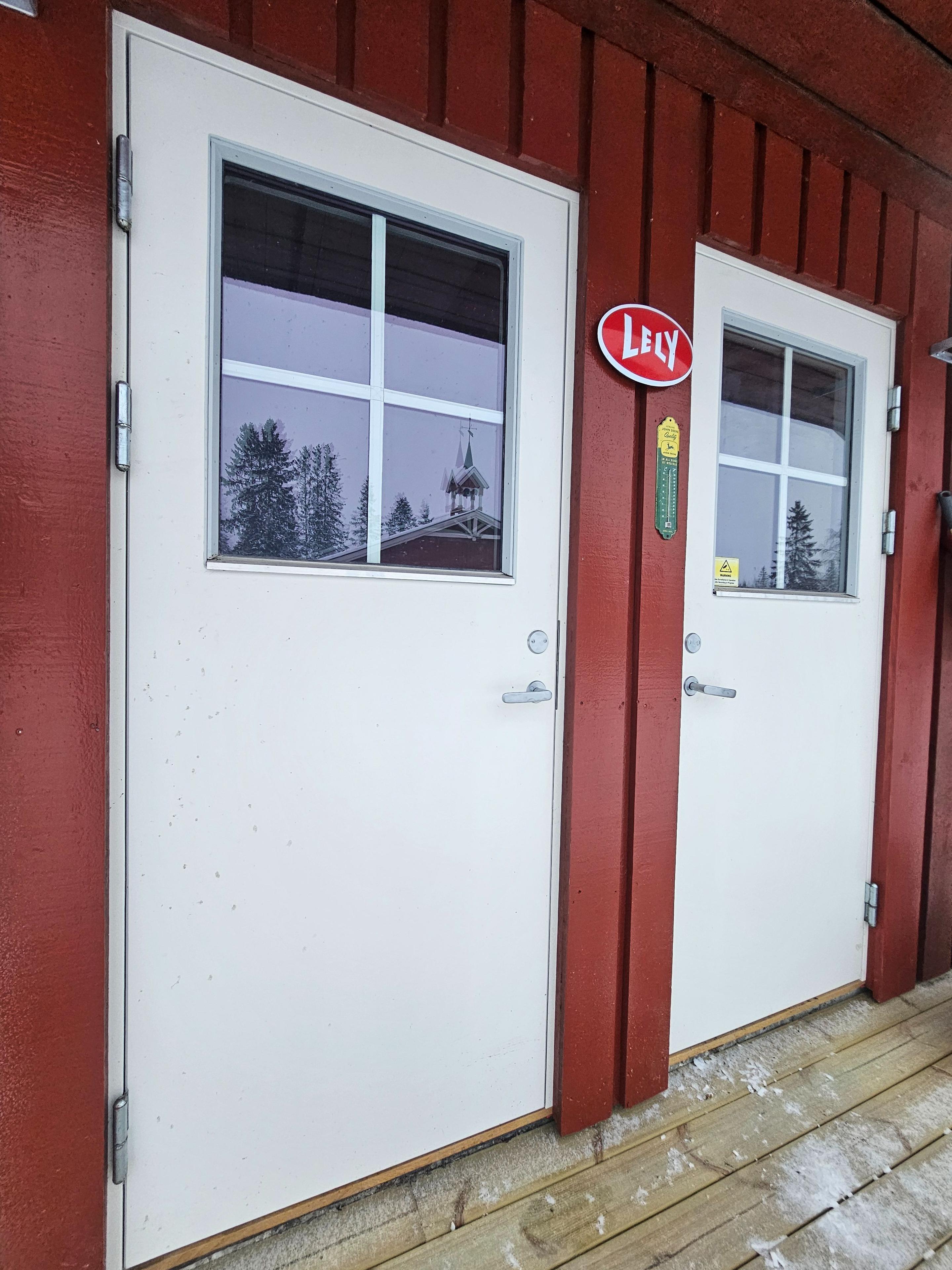 To dører på en rød hytte med snø på bakken og trær i bakgrunnen.