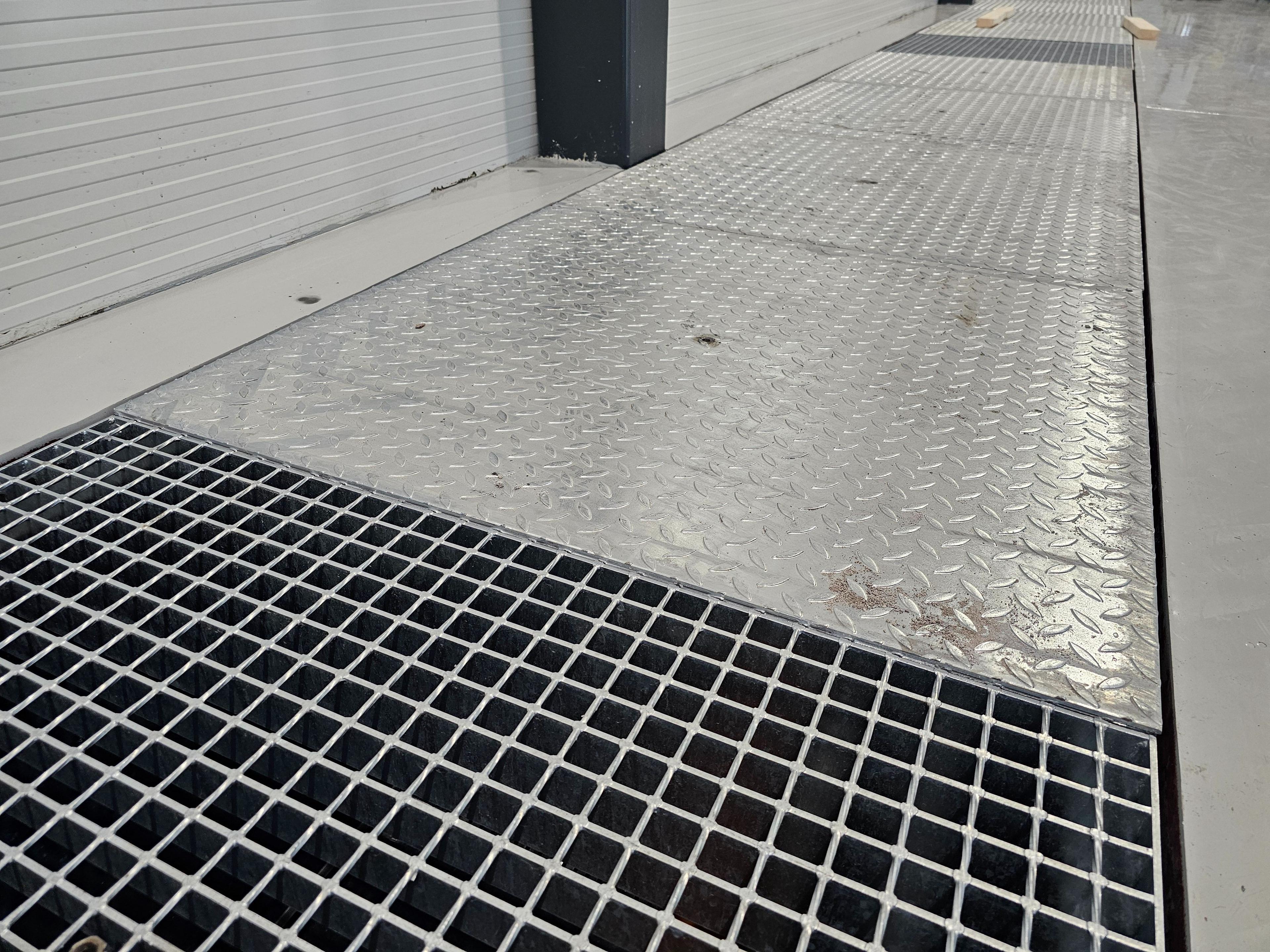 Et metallgitter på en gangvei med rutenett og skinnende overflate, plassert mellom bygninger.