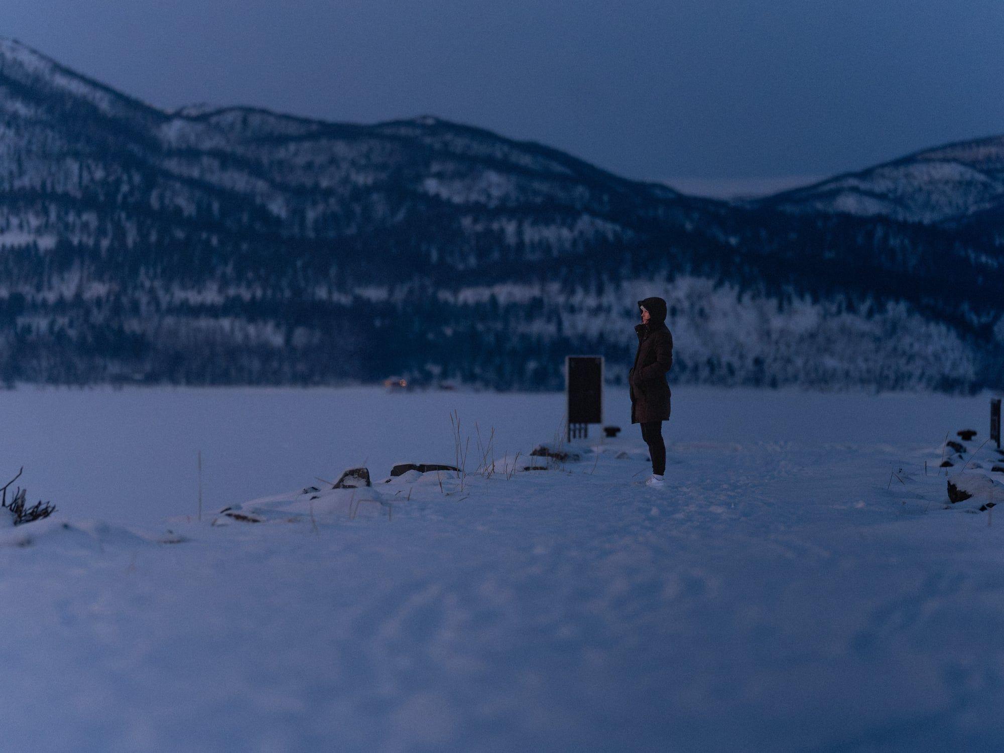 En person står på en snødekt strand ved en innsjø, med fjell i bakgrunnen under en stjerneklar himmel.