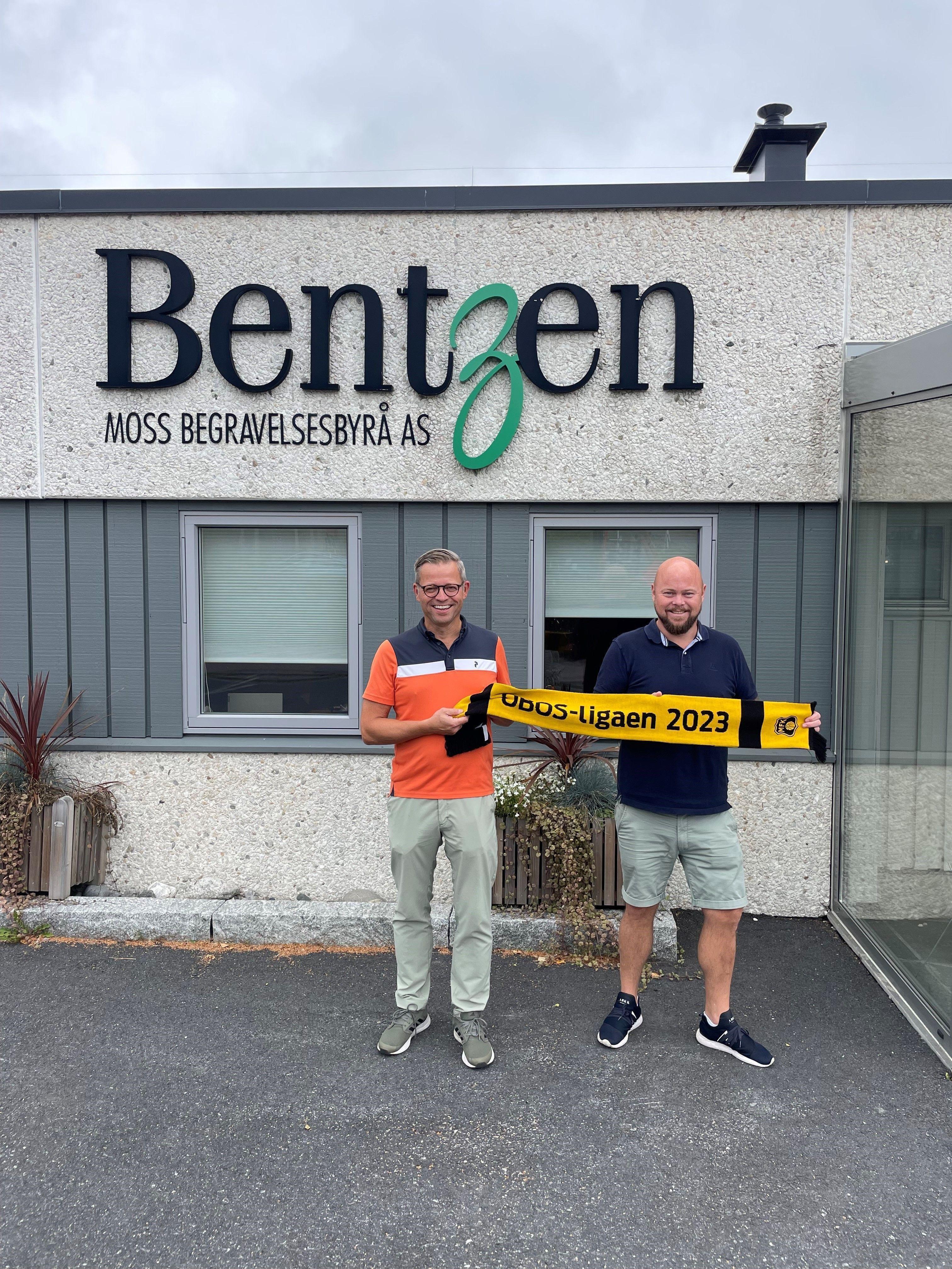 To personer står foran Bentzen Moss begravelsesbyrå AS med en banner for "Moss-Ligaen 2023".
