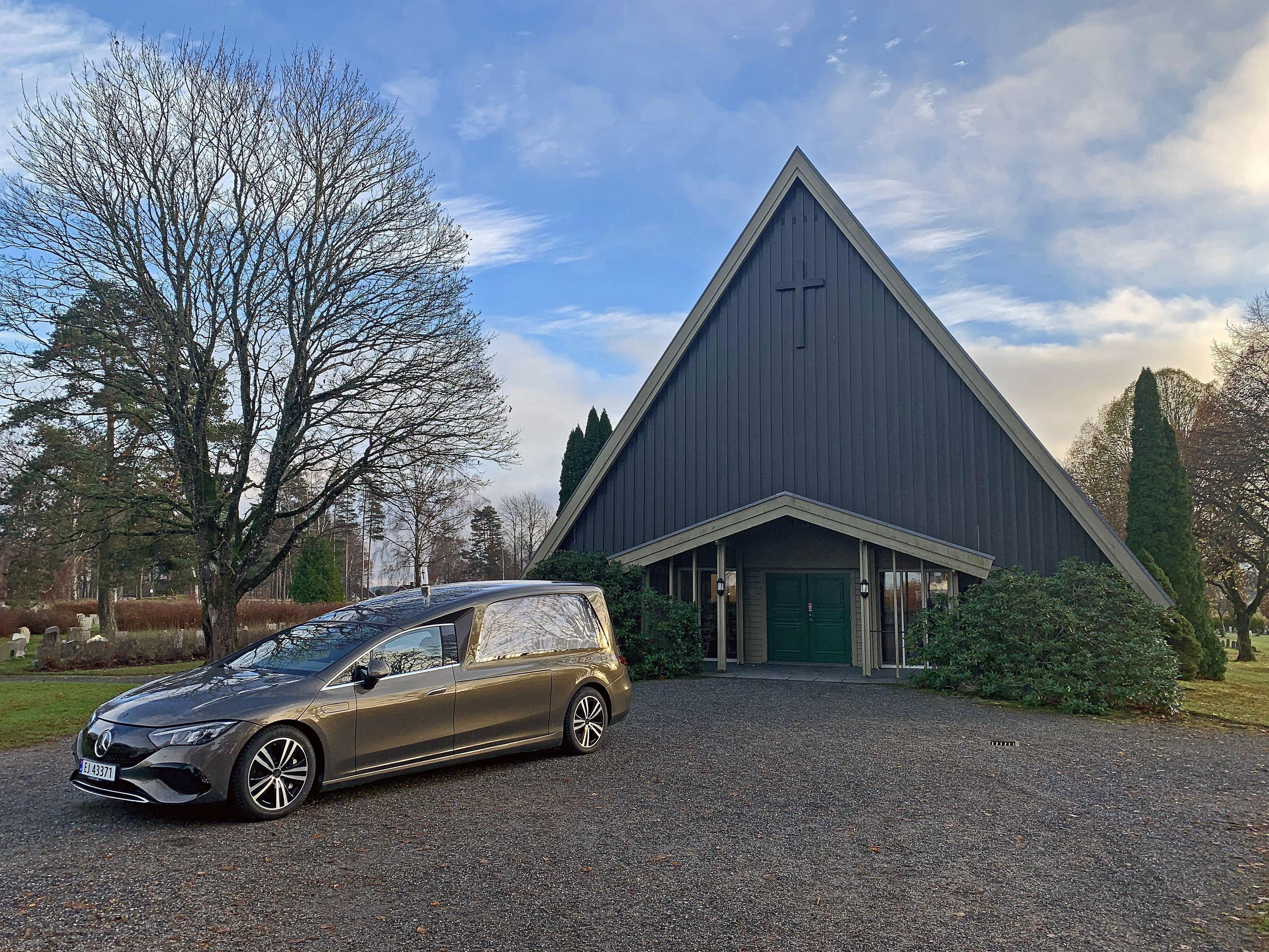 En grå bil parkert foran en moderne kirke med trekantet tak og grønne busker rundt.