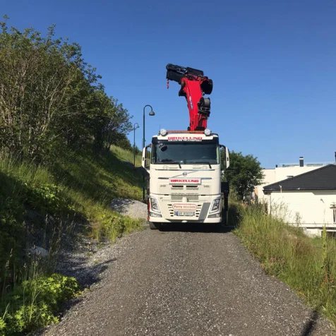 En lastebil med kran står på en grusvei omgitt av grønt gress og busker under en klar blå himmel.