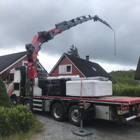 En lastebil med kran som løfter pakker foran et hus under en grå himmel.