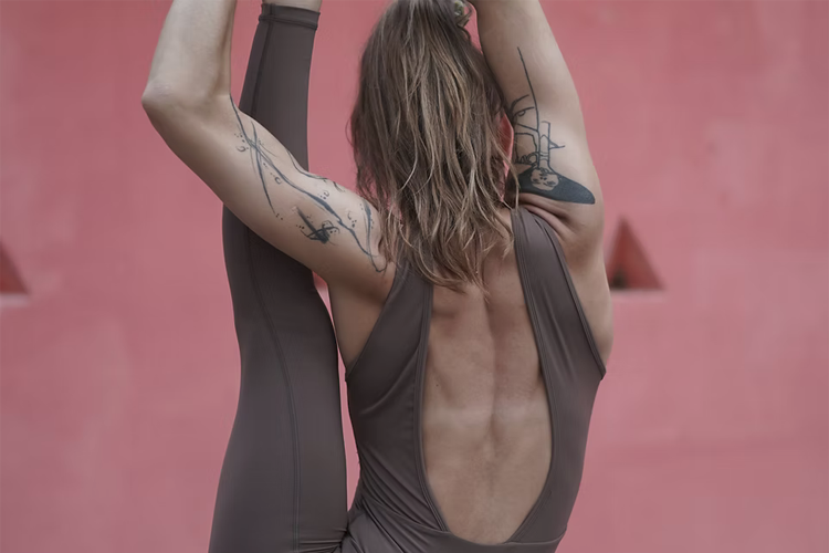 Kvinne i yogaøvelse med tatoveringer, iført brun treningstights, mot en rosa bakgrunn.