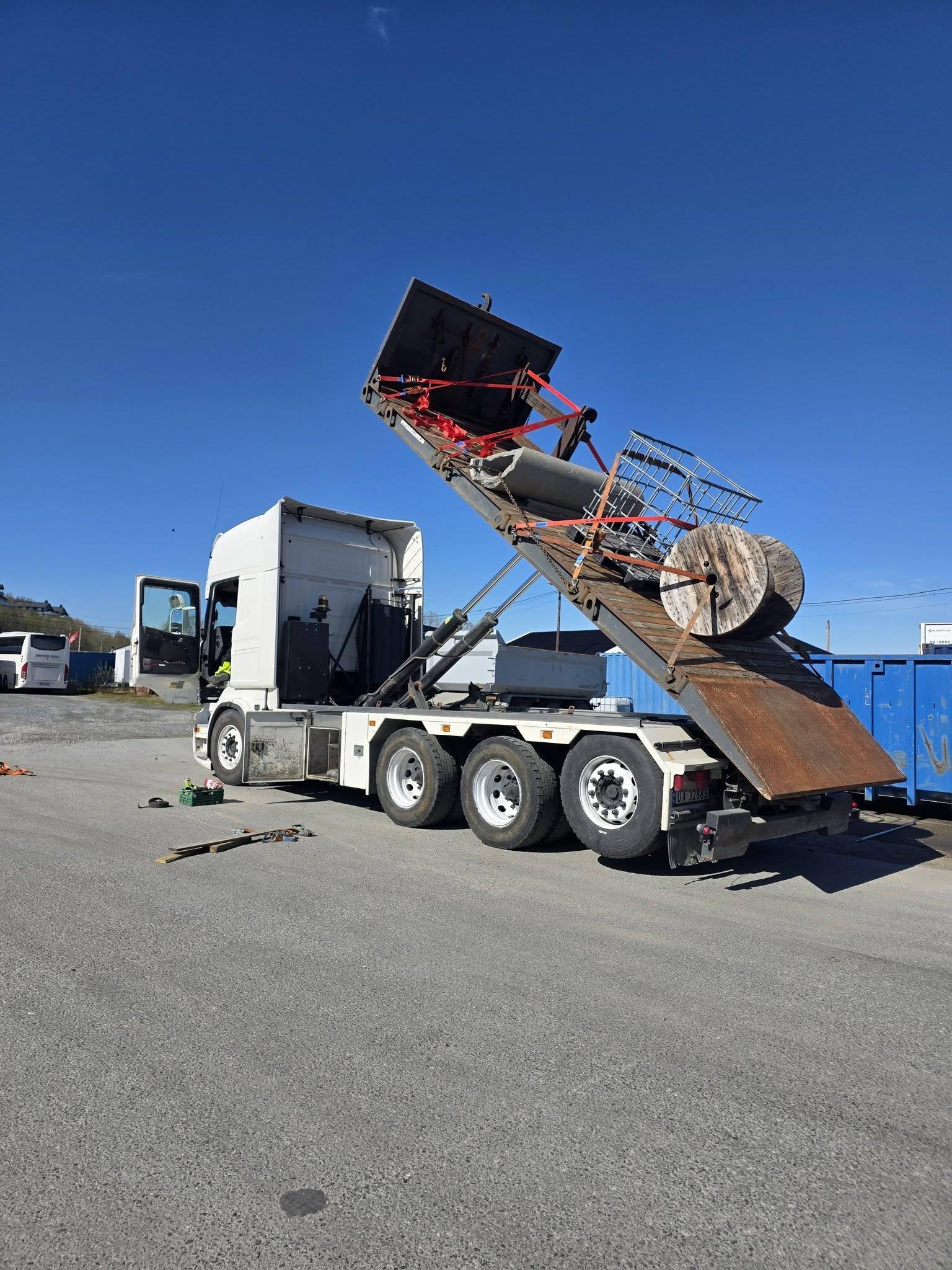 En lastebil med en hevet bakluke som laster av materialer på en byggeplass under klar himmel.