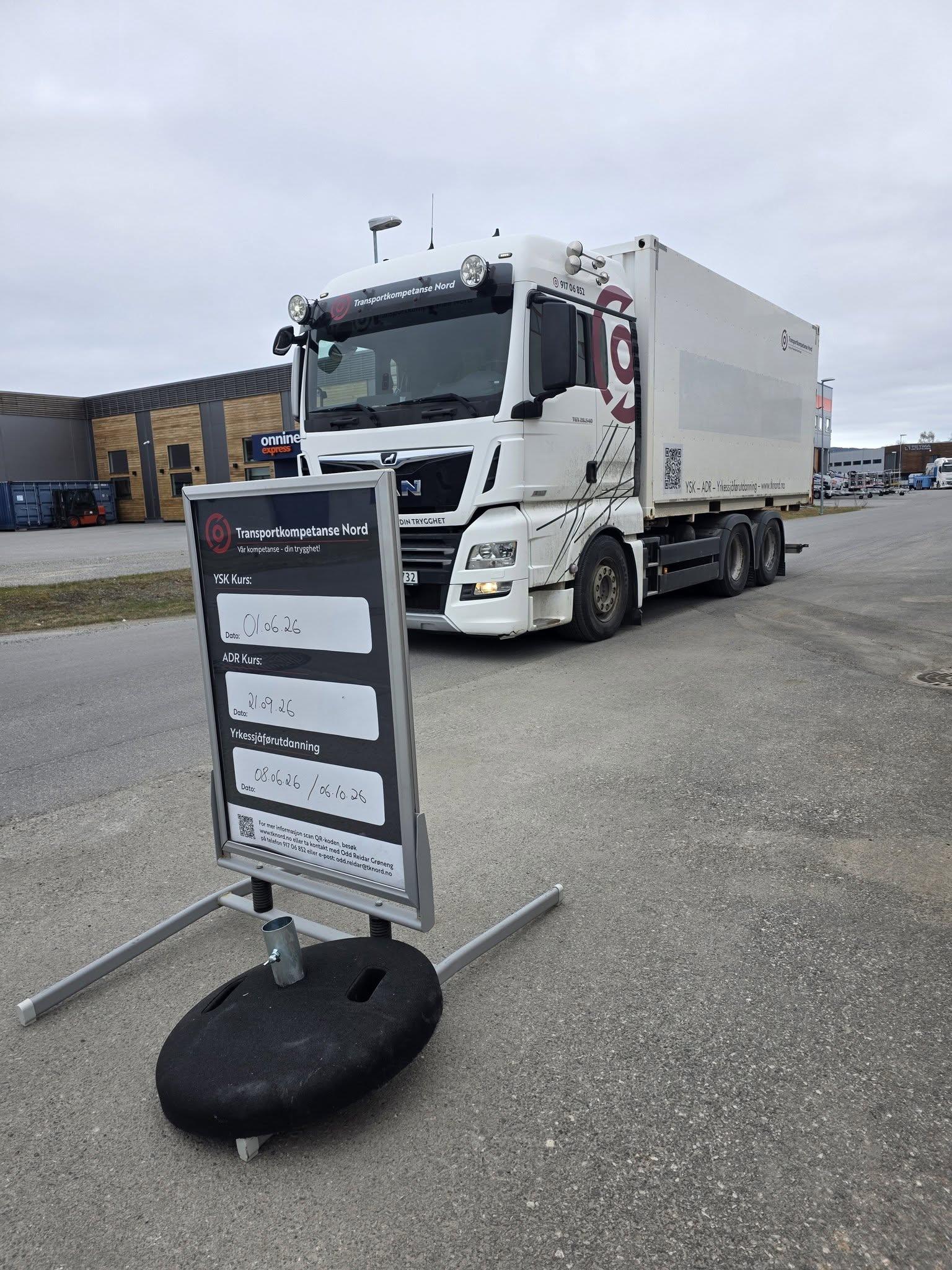 Lastebil med tilhenger parkert ved et skilt som viser informasjon om området. Grå himmel i bakgrunnen.