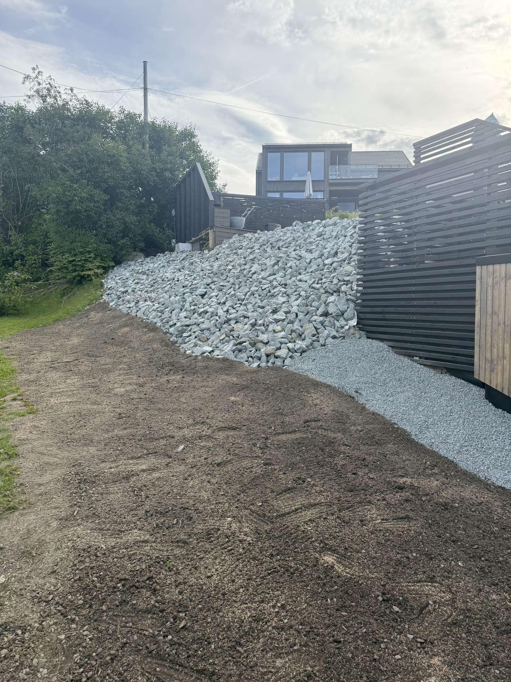 Gruslagt skråning med stein og moderne hus i bakgrunnen, omgitt av grønt landskap.