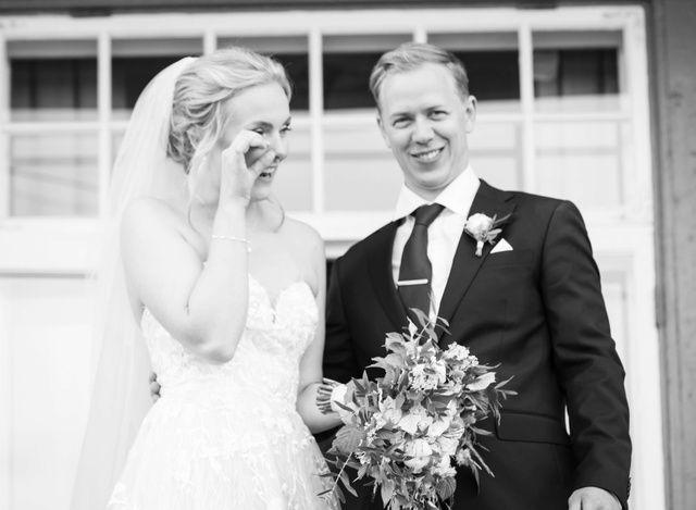 Brudeparet smiler sammen, bruden i hvit kjole med slør og brudgommen i svart dress, holder en bukett blomster.