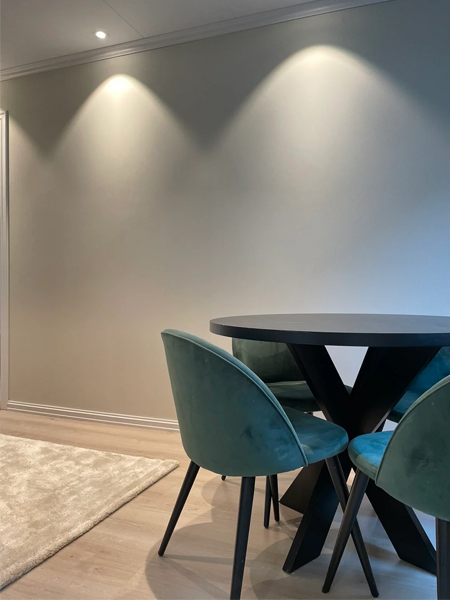Moderne spisebord med svarte ben og grønne stoler, plassert på et lyst rom med vegglys og teppe.