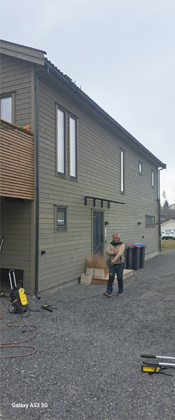 En mann står foran en moderne, grå bygning med store vinduer og en terrasse. Søppelbøtter er synlige i bakgrunnen.