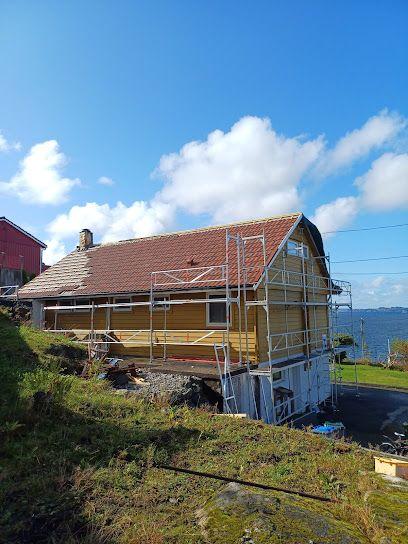 Hus med stillas og takrenovering, beliggende ved sjøen under en blå himmel med skyer.