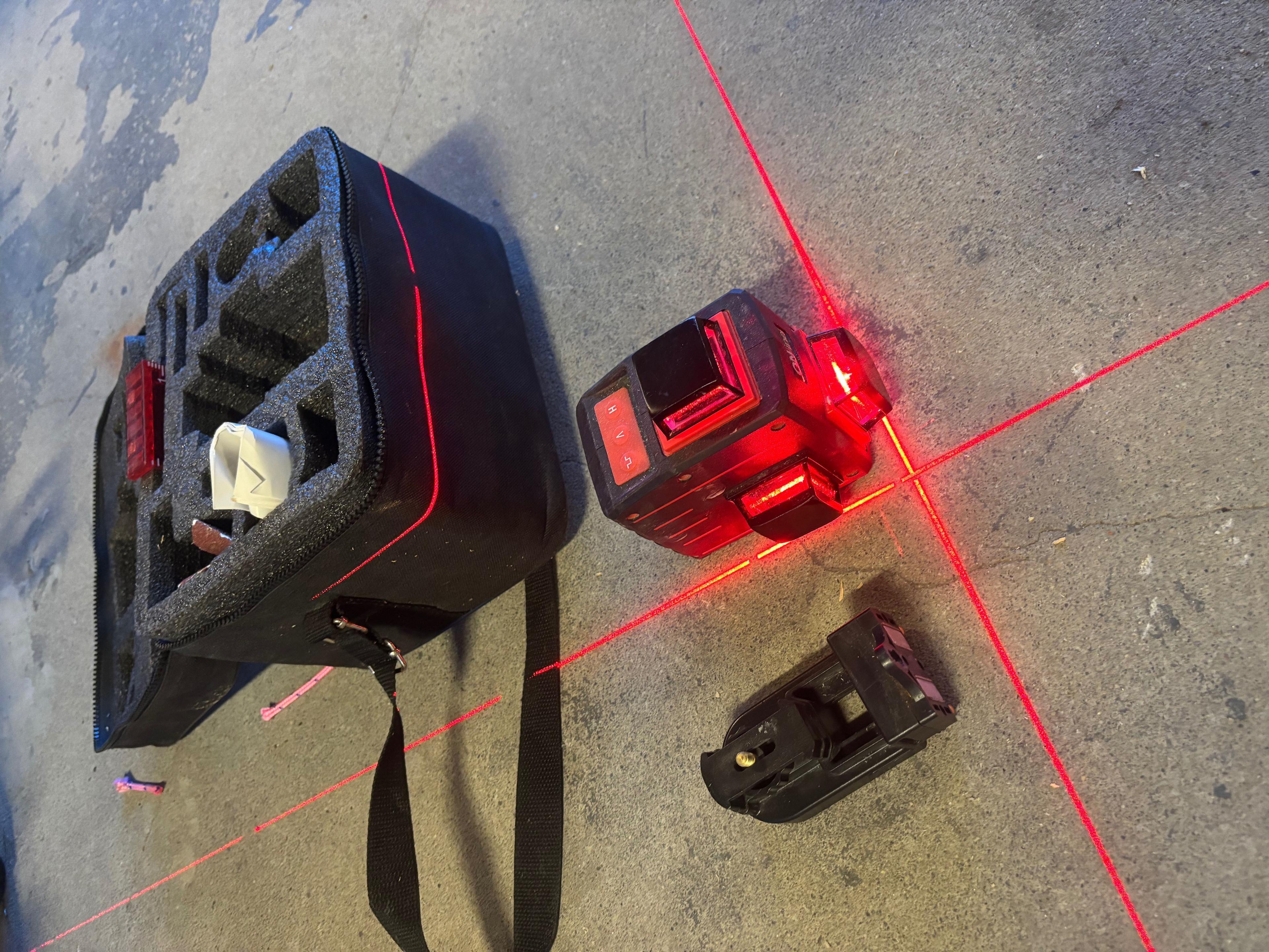 Laserverktøy med rød laserlinje, en bæreveske og en festebrakett på betonggulv.