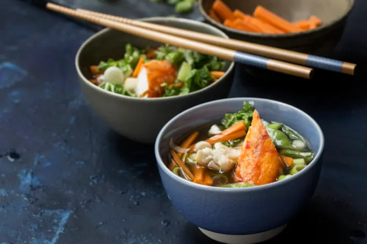 En bolle med suppe med grønnsaker og fisk, ved siden av en bolle med salat og spisepinner.