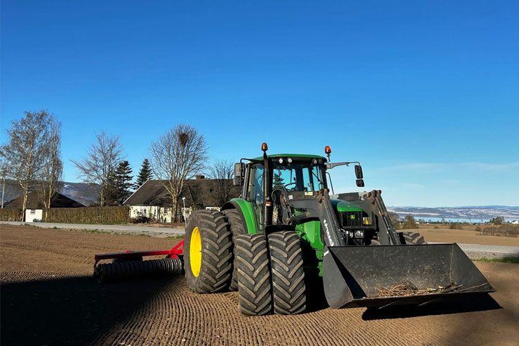 En grønn traktor med stor skuff står på en åker under en klar blå himmel.