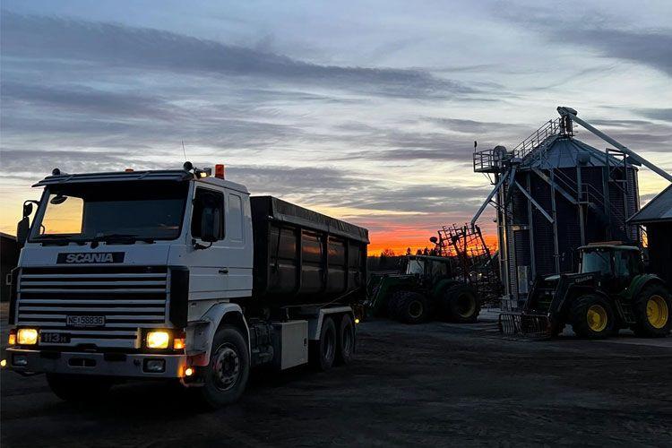 En lastebil med tilhenger står foran en sil ved solnedgang, med traktorer i bakgrunnen.