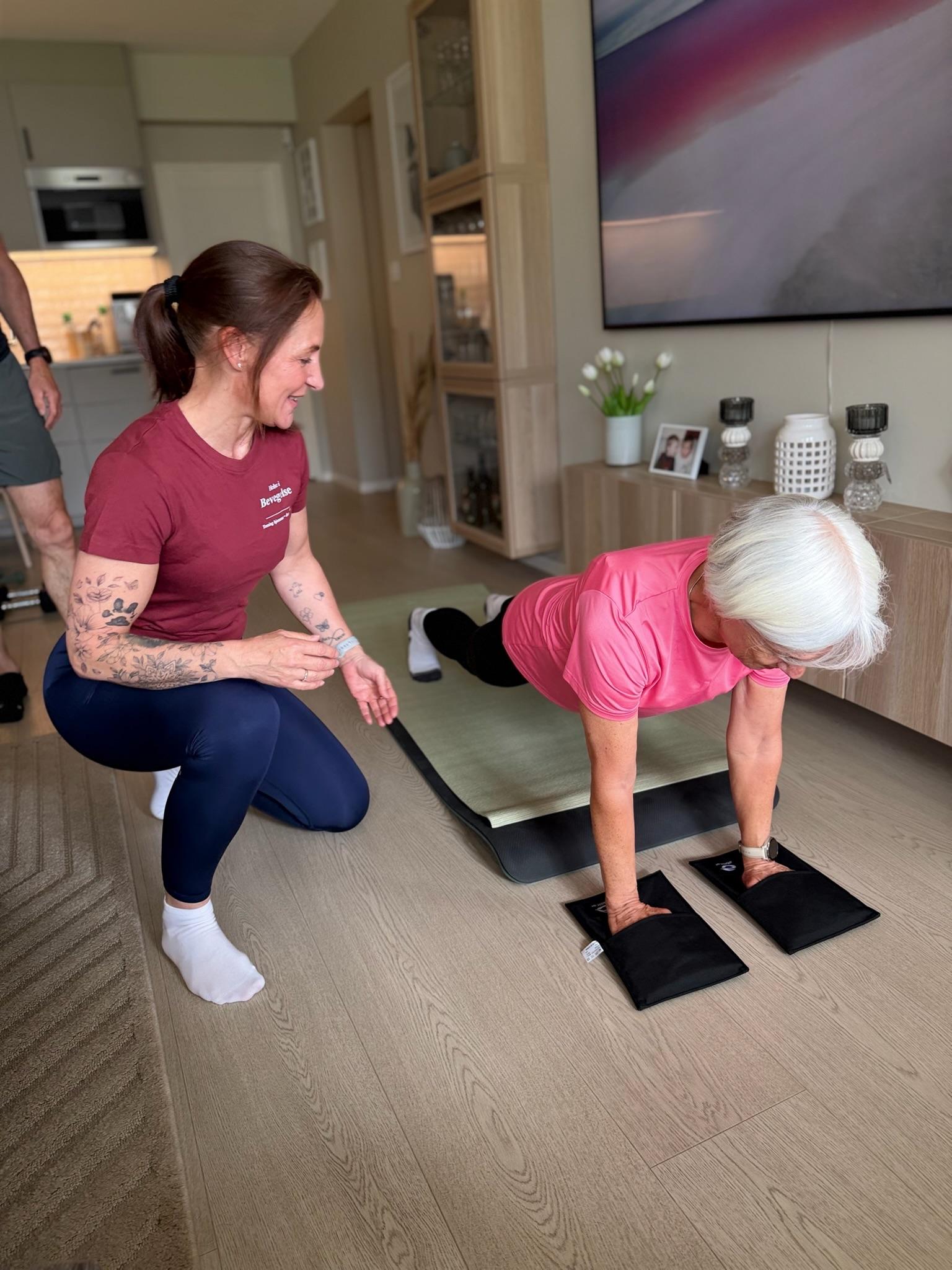 En kvinne trener med en eldre dame som gjør push-ups på en matte, mens en instruktør veileder henne.