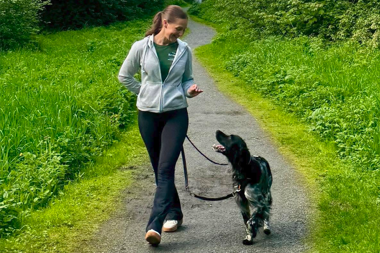 En kvinne går på en sti med en hund i bånd, omgitt av grønt gress og trær.