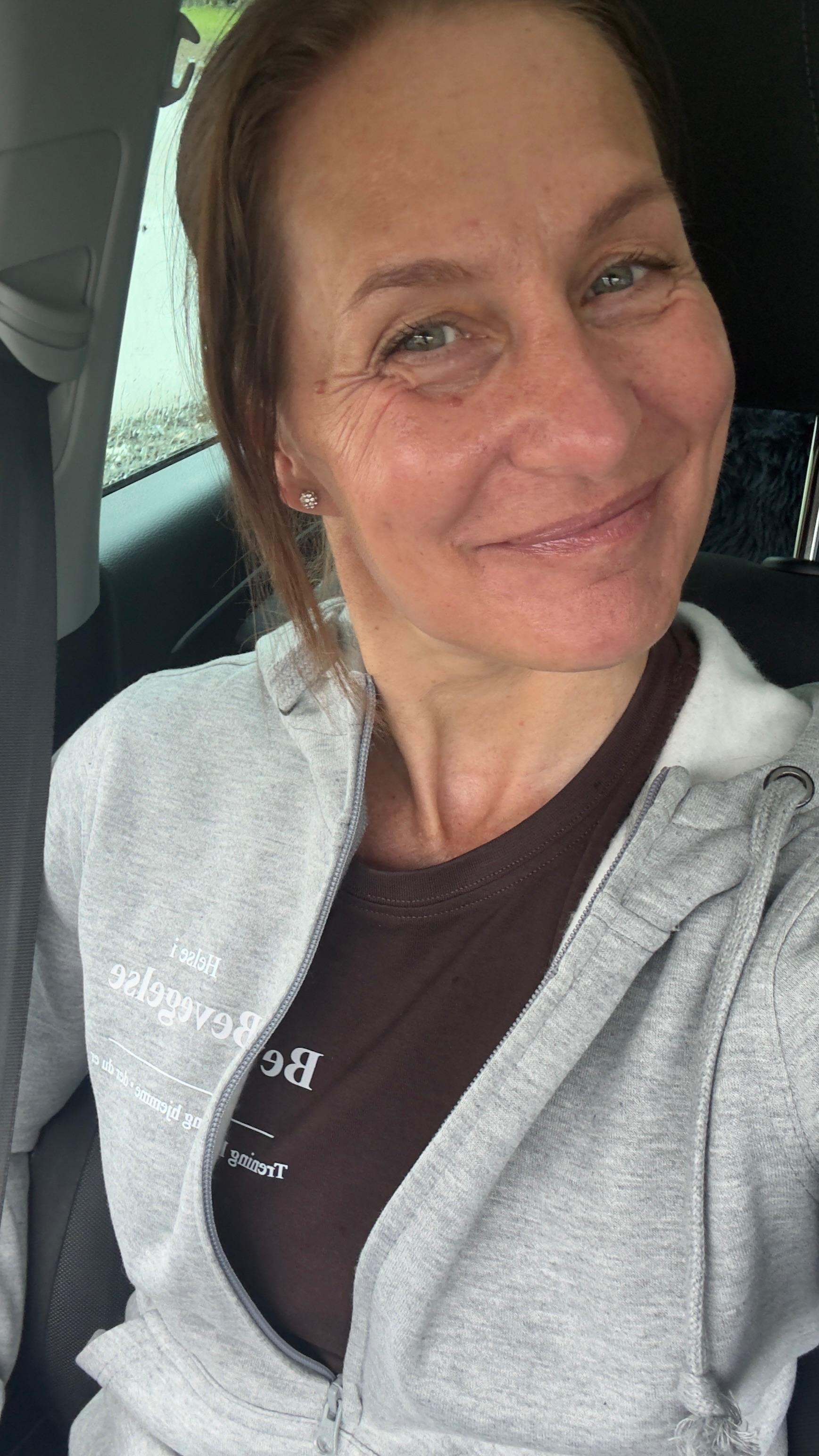 Kvinne smiler i bilen, iført en grå hettegenser og en brun t-skjorte.