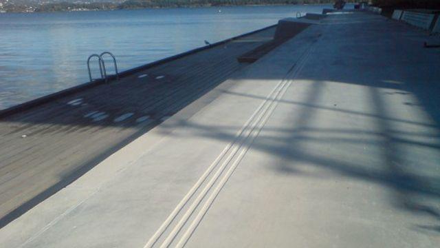 Bred betongpromenade ved vannkanten med skygger og en svømmerampe i bakgrunnen.