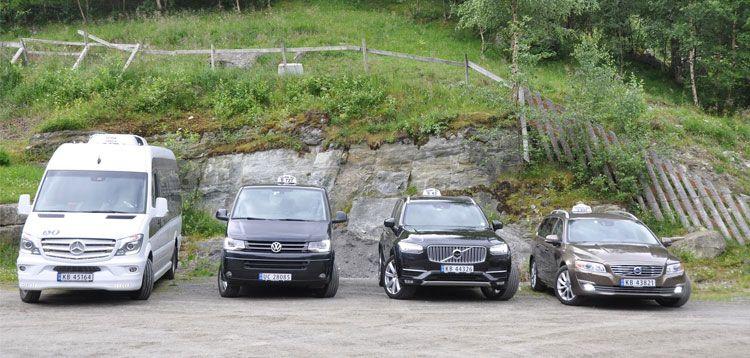 Fire biler parkert på en grusplass, inkludert en hvit minibuss og tre svarte personbiler.