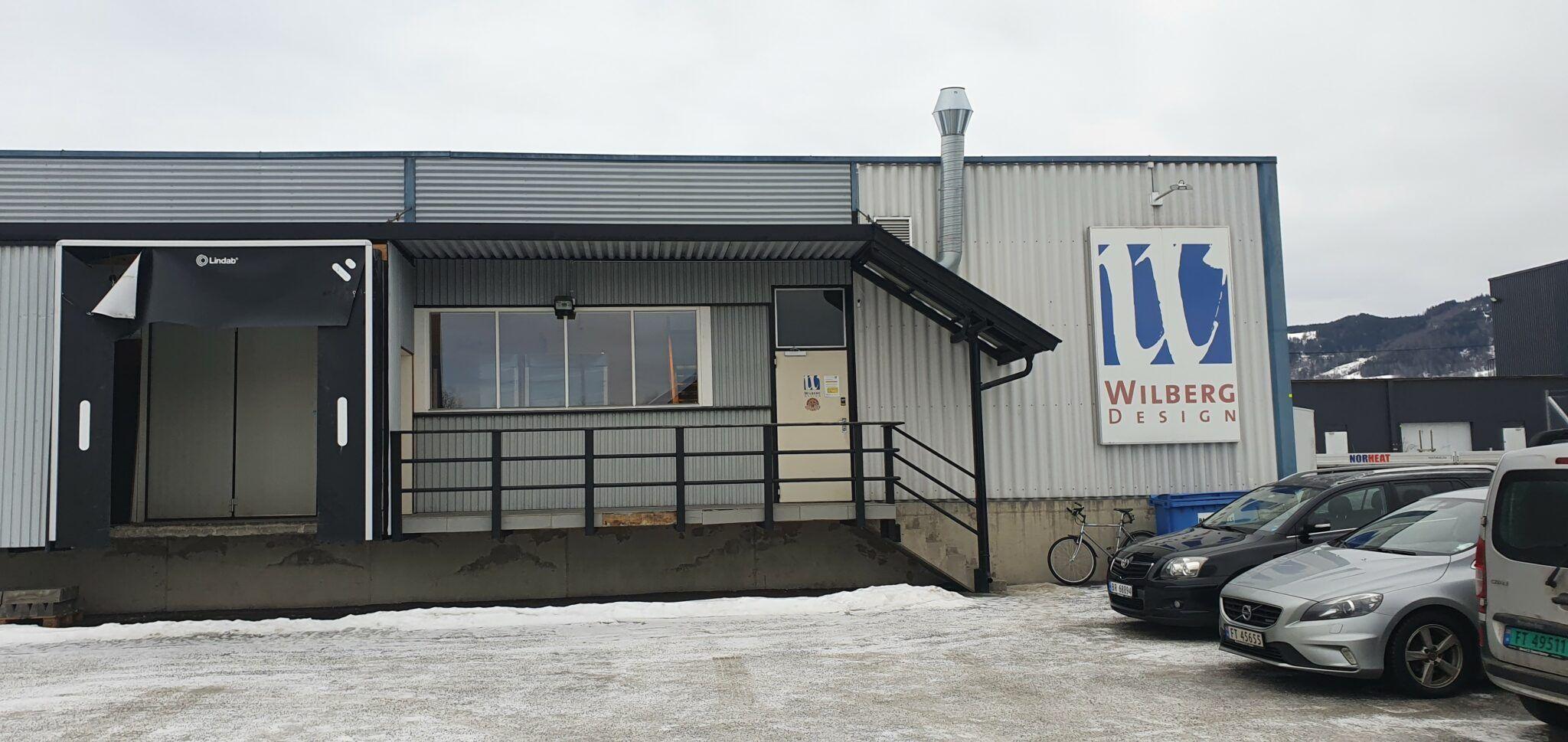 Bygning med Wilberg-logo, parkerte biler og snø på bakken, under en grå himmel.