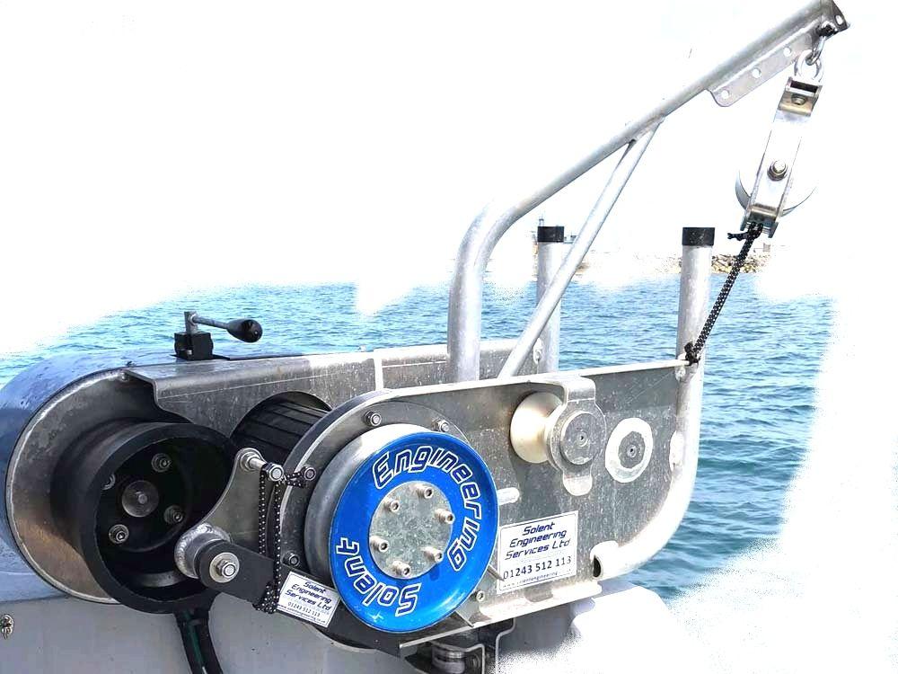 En båtwinch med metallkonstruksjon og blå spole, plassert på en båt med havet i bakgrunnen.