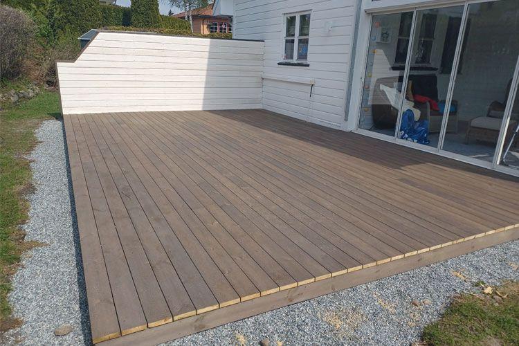 Terrasse i tre med grå stein rundt, ved siden av et hus med store vinduer.