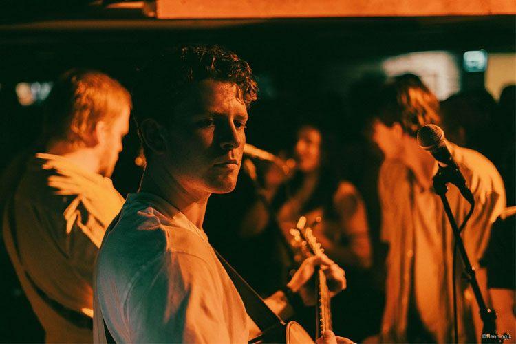 En musiker med gitar ser mot kameraet mens han spiller på en konsert med publikum i bakgrunnen.
