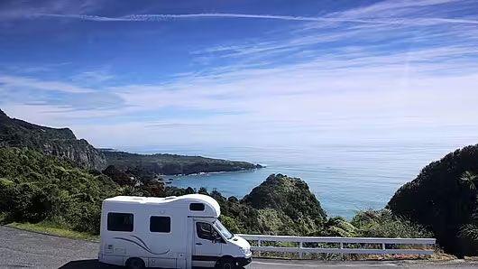 En campingvogn står ved veien med utsikt over havet og fjellene under en klar blå himmel.