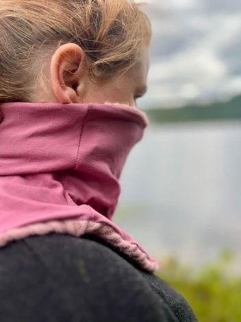 En person med rosa halsvarmer står ved vannet, med ryggen til kameraet og naturen i bakgrunnen.
