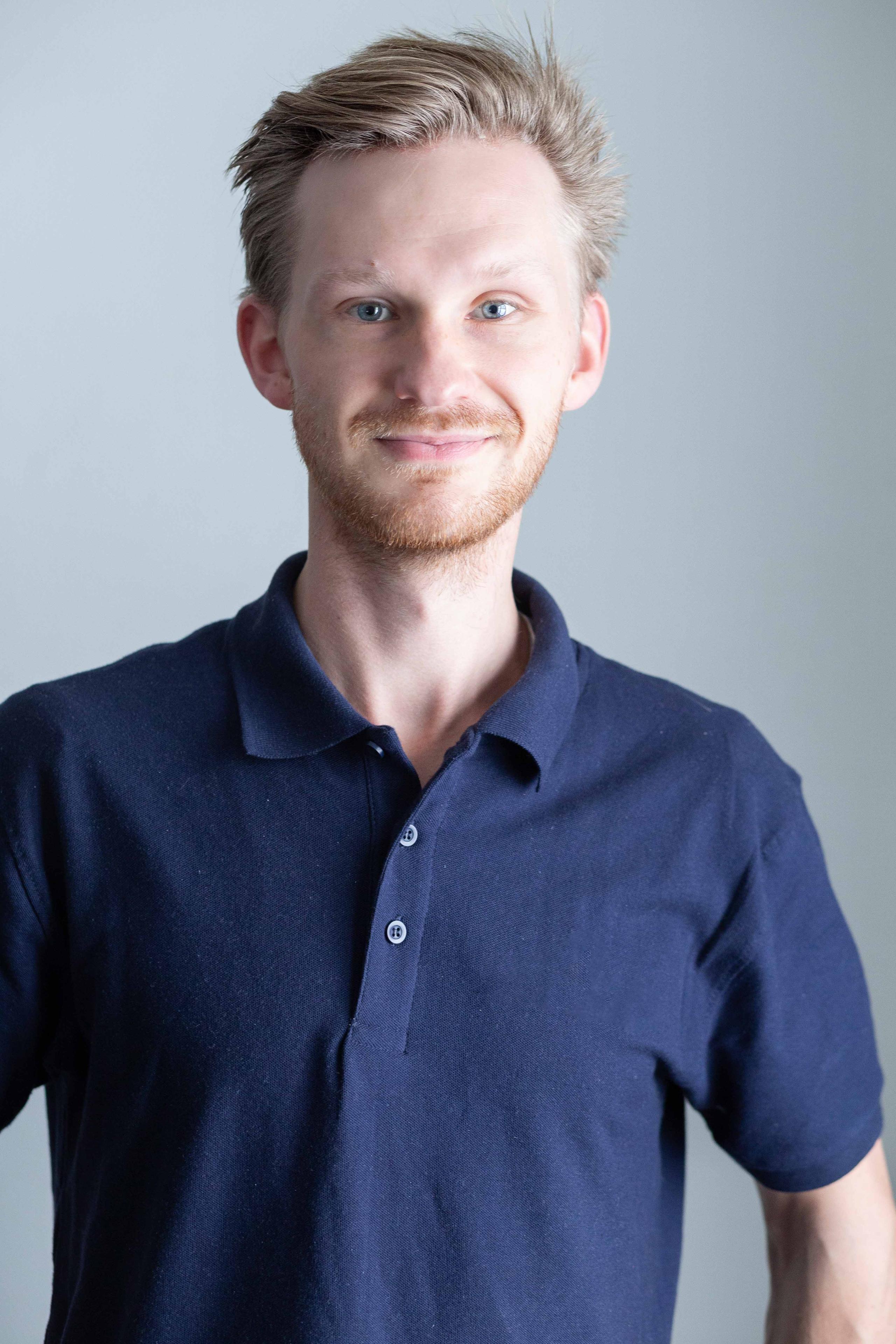 En smilende mann med lyst hår iført en blå polo skjorte, står foran en lys bakgrunn.