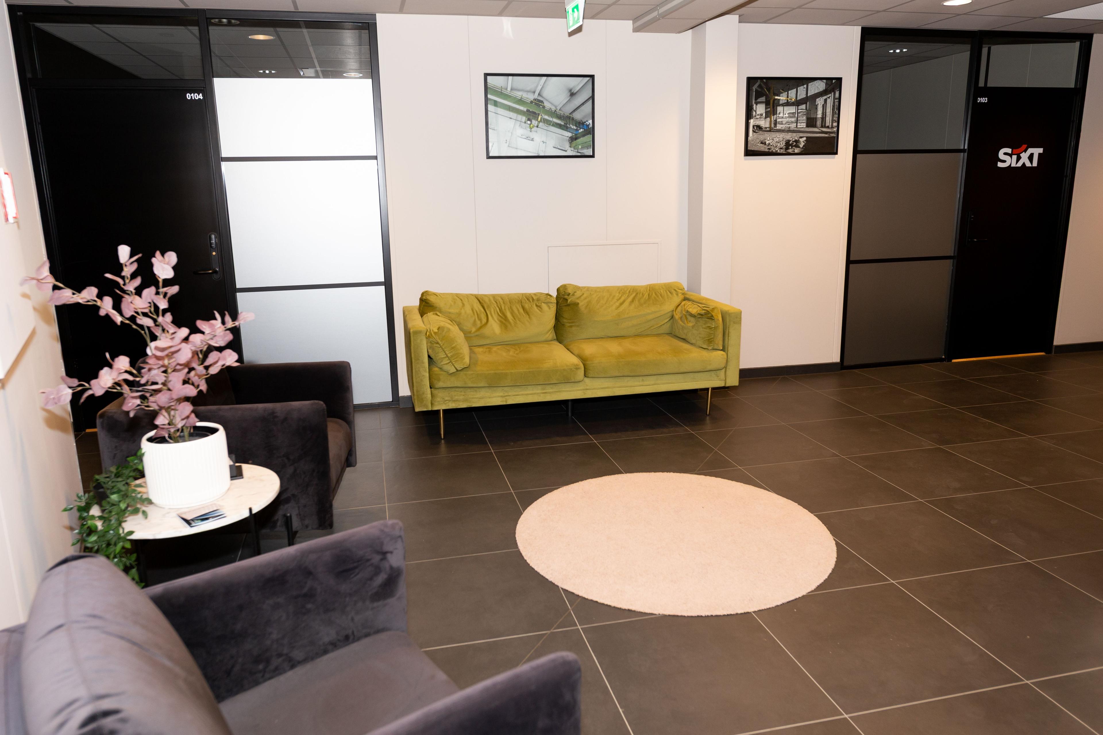 Moderne kontorlokale med grønn sofa, stoler og planter, samt glassdører til møterom.