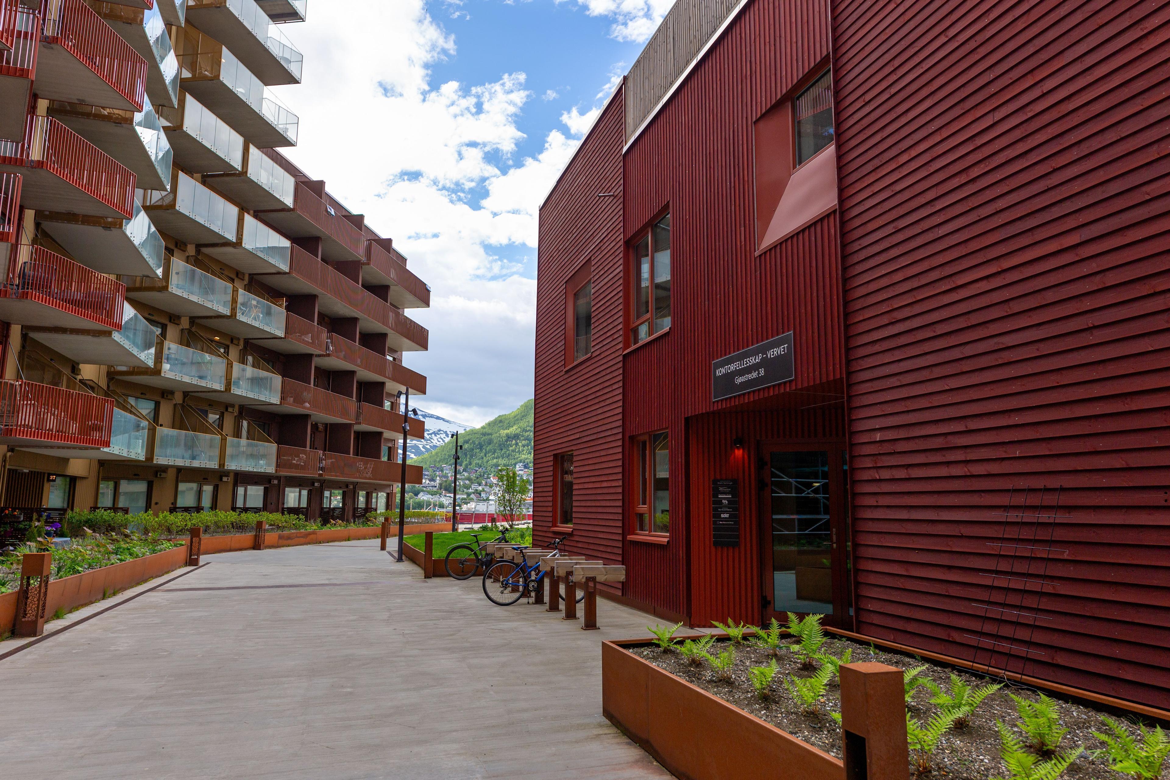 Moderne bygninger med rød fasade og grønt landskap i en urban gate. Sykkelparkering synlig.