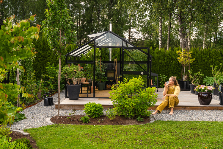 En kvinne sitter på terrassen foran et moderne drivhus omgitt av grønne planter og blomster i hagen.