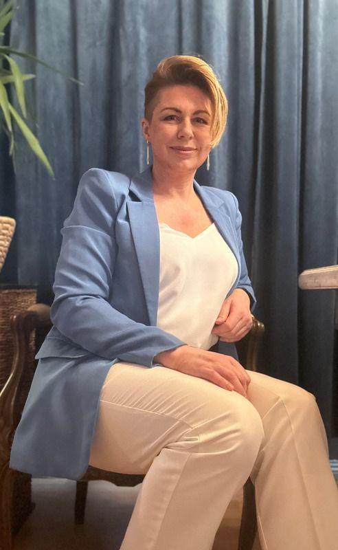 Kvinne med kort hår iført blå blazer og hvit topp, sitter i en stol foran en mørk bakgrunn.