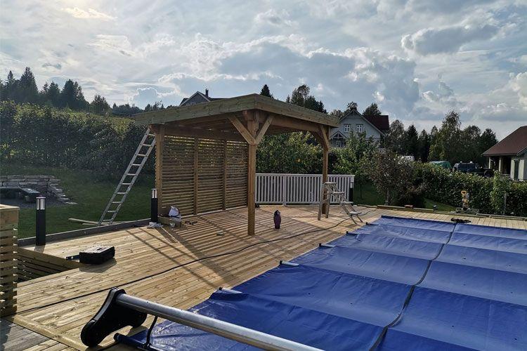 Uteområde med en trepergola, svømmebasseng med blå dekke og en stige i bakgrunnen, omgitt av grønne planter.