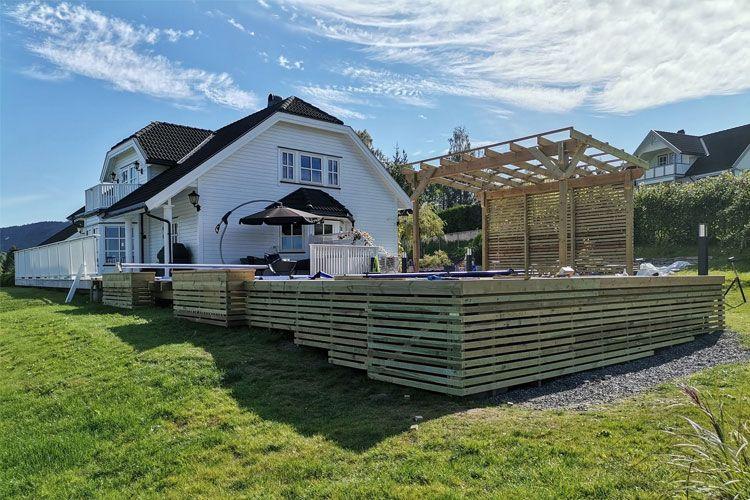 Moderne hus med stor terrasse og soldekk, omgitt av grønt gress og blå himmel.
