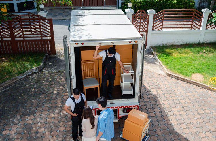 To personer laster møbler inn i en flyttebil, mens en annen person står ved siden av og ser på.