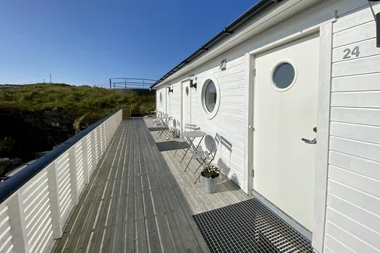 En moderne hytte med rund vindu, terrasse og stoler, omgitt av grønt landskap under en klar blå himmel.