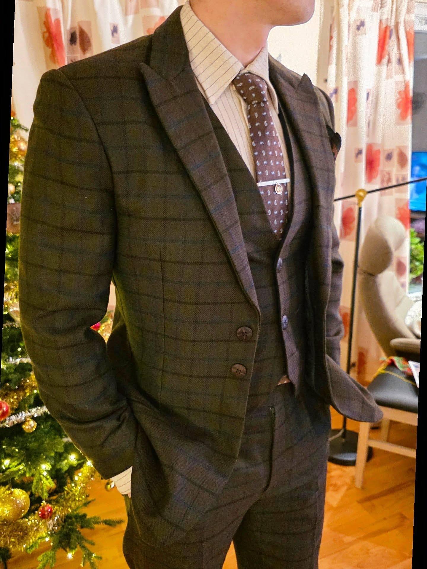 En mann i en rutete dress med vest og slips, står foran et juletre i et koselig rom.