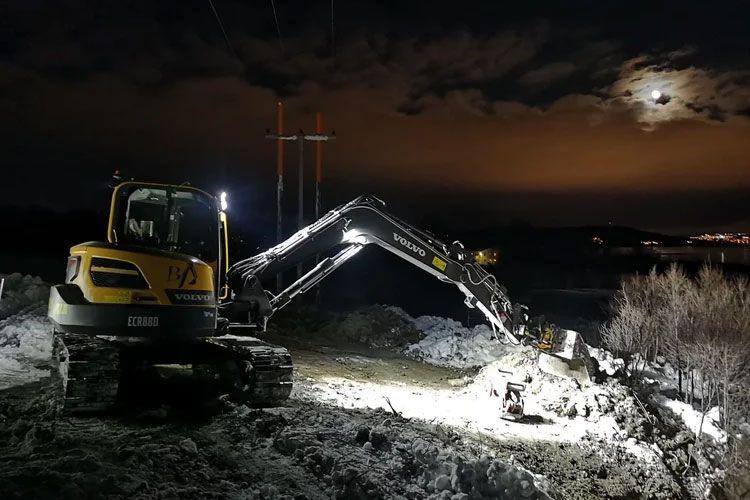 Gravemaskin arbeider i mørket med lys, ved en snødekt vei under måneskinn.