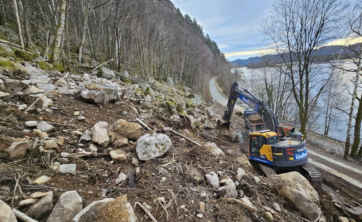 Gravemaskin som arbeider med stein og trær langs en vei ved en innsjø.