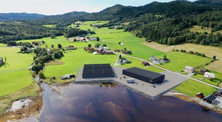 Luftbilde av en landsby med grønne åser, bygninger og en stor svart bygning ved vannkanten.