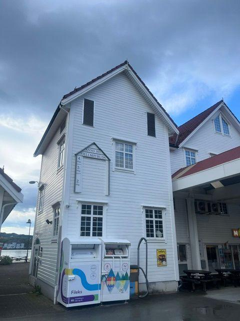 Hvit bygning med skråtak og vinduer, med en kiosk foran og mørke skyer i bakgrunnen.