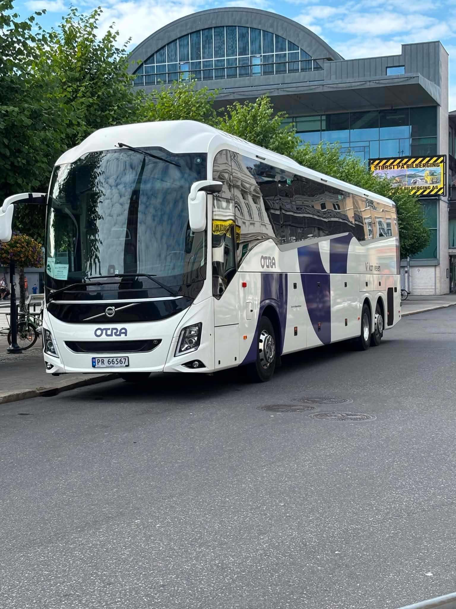 En moderne turistbuss parkert ved en bygning med glassfasade og trær i bakgrunnen.