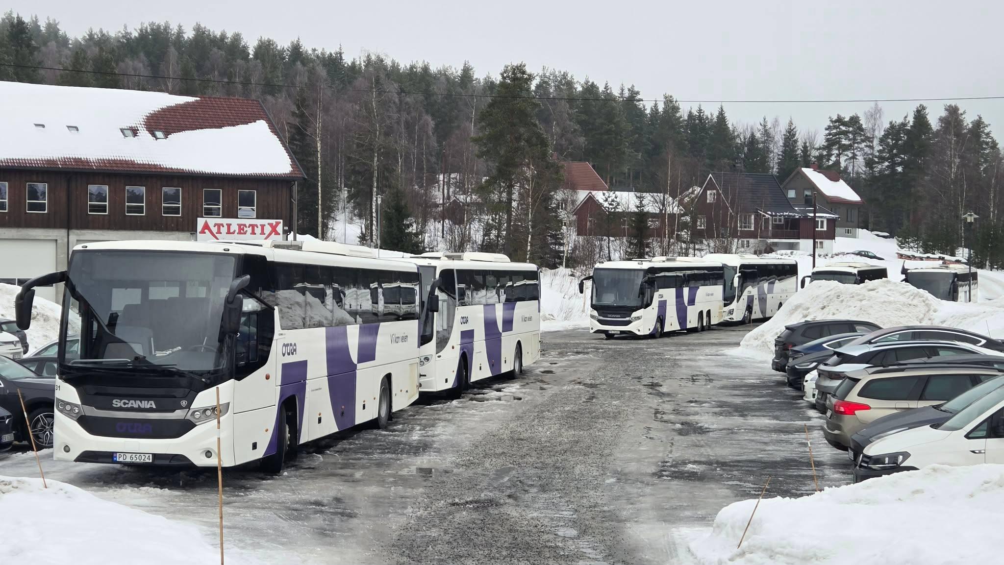 Flere hvite busser parkert på en snødekt parkeringsplass med trær og bygninger i bakgrunnen.